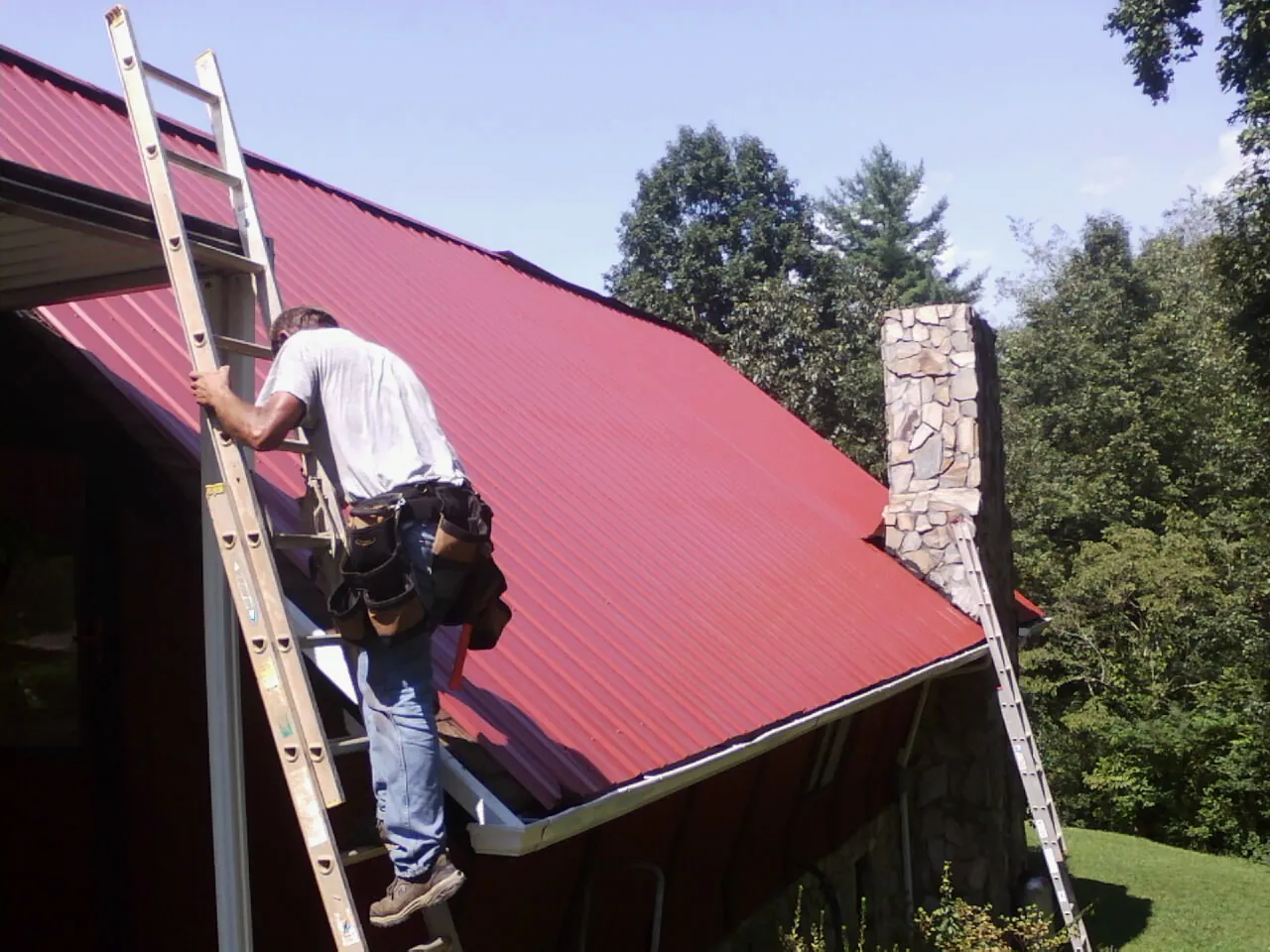Roofer on a ladder beside a red metal roof and stone chimney — Soffit & Fascia Repair by Roofing Brothers in Allenhurst